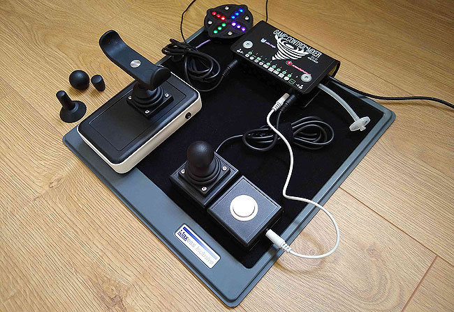 A complex Game Control Mixer accessible gaming set-up with two chunky joysticks, one with a MERU red U cow bar fitted. Two Sanwa switches and a small Atec ultra light blue switch. A microphone, PS4 controller and Titan Two adapter on a velcro tray.