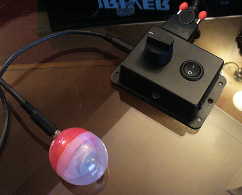 Image of a black adapted Xbox 360 joypad, connected via chunky Lindy SVGA cables to a box with a dial, on/off switch and two sockets. One socket for a switch, the other for a Vibro Capsule (Red and clear one with motor inside attached). On the other side an external thumbstick in a small sloping black case is attached.