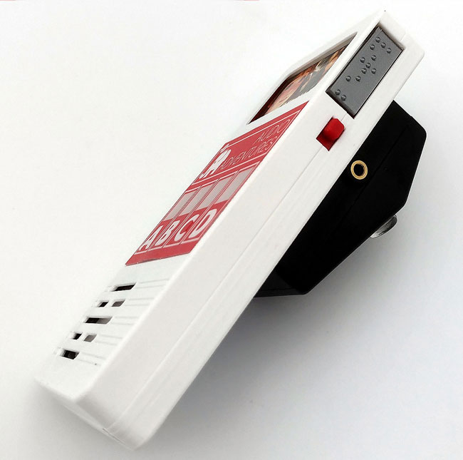 Side view with Braille game cartridge label at the top. Red on/off switch. Black angled switch box at the back with rubber foot on show.