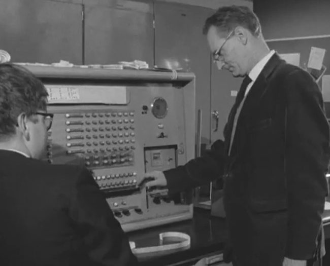 Black and white photo of a National-Eliott 405 computer from the 1950s being used by a teacher playing their reaction tester game, monitored by a pupil. Via BBC's Tomorrow World in 1969.