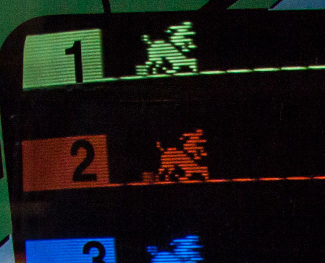 Atari Steeplechase gameplay photo. Seven individually coloured rows, each with a uniformly broken line representing the ground.   From left to right, a block to illuminate an number. Then a horse and jockey. Some block hurdles then at the end chequered blocks to represent the finish line.  The colour and numbers come from a plastic overlay.