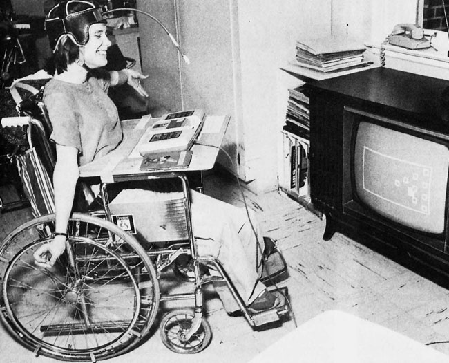 Smiling young woman in a wheelchair playing a very basic black and white television bowling game using a head mounted pointing device to push down on the keys on a RCA Studio II games console.