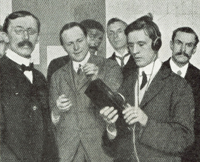 An academic demonstration. From left to right in front of a table of electronic devices:  Fournier D'Albe in bow-tie and suit stares towards the camera with hand protectively on the Exploring Optophone box.  Mr. Arthur Burrows, director of programmes of the British Broadcasting Company, holds a lit match.   Mr. B, wearing headset, points the exploring Optophone at the burning match.  Behind this three other gentlemen stand at awkward angles to make sure they are also in the photo.