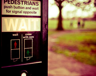 One Switch 100. UK Pedestrian crossing control, with push-button and WAIT instructions. In the distance three blurry figures walking across a filed towards the push-button.